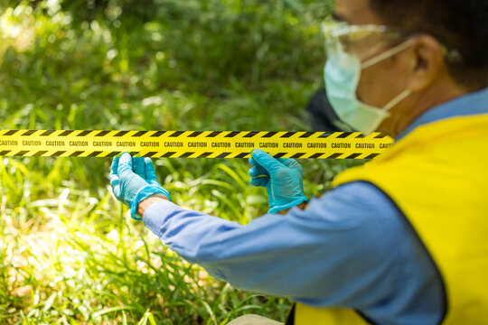 Masked field worker in protective clothing and blue gloves examines evidence behind yellow caution tape in a sunlit green outdoor area.