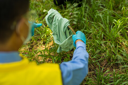 Worker in a yellow vest and blue gloves picks up a green discarded garment from tall grass during an outdoor cleanup.