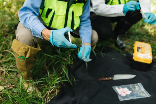 Forensic investigators in protective gloves collect and document evidence outdoors on a black evidence sheet with tools, bags, and a yellow case.