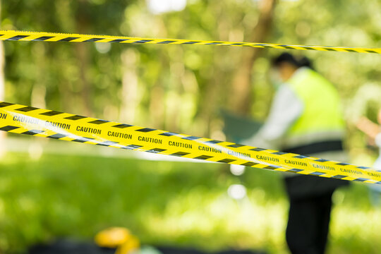 Yellow caution tape stretches across a sunny wooded outdoor area, with a blurred safety worker in a high visibility vest behind it and soft green foliage in the background.