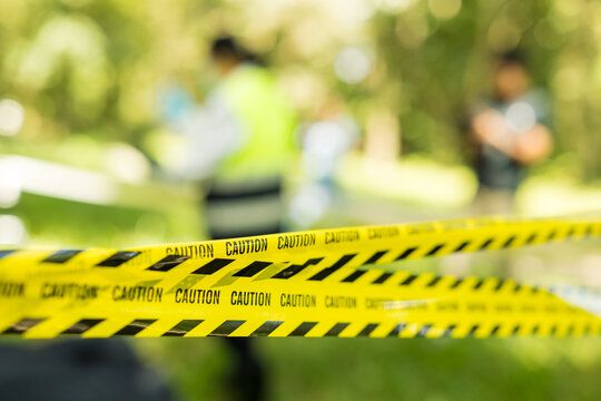 Yellow and black caution tape blocks an outdoor area while blurred people stand behind it, suggesting a police investigation or safety emergency.