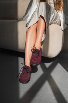 Close-up of woman seated on sofa with crossed legs, burgundy lace-up shoes, and flowing beige dress under strong natural sunlight with bold shadows and warm mood.