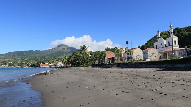 St Pierre beach showing sailboats moored in the bay with historic buildings along the coastline and the active Mount Pelee volcano,rising majestically in the background under a blue sky in Martinique