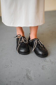 Black leather lace-up shoes with white midi dress, close-up of feet on gray concrete floor, minimalist urban fashion detail with neat lacing and neutral lighting