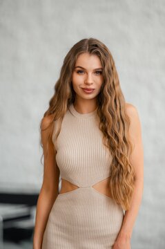 Close up portrait of confident young woman with long wavy brown hair wearing beige ribbed sleeveless dress with waist cutout in soft neutral gray studio lighting