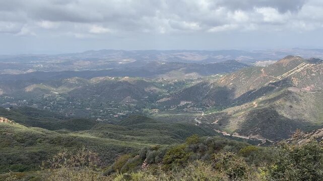 Sunlight and cloud shadows drift over green canyons and rocky ridges of the Santa Monica Mountains seen from Lois Ewen Overlook in Topanga, California, with homes scattered in the valley below.