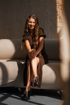 Smiling woman in brown knit dress relaxing on cream outdoor sofa, long wavy hair and black lace-up shoes, warm sunlight and soft shadows in sunny garden, calm mood