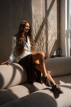 Woman longhair brunette in white blazer belt sits on cream sofa in sunlit modern interior by window with textured wall and decorative branches, relaxed stylish pose