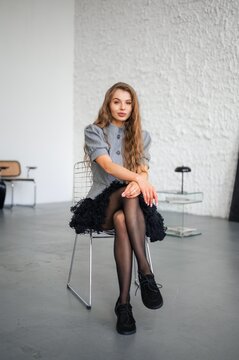 Stylish woman seated on metal wire chair in minimalist studio, long wavy hair and soft makeup, gray puff-sleeve top, black skirt and lace shoes, textured white wall background