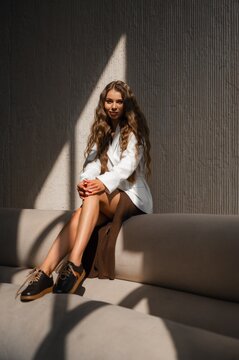 Portrait of stylish woman seated on concrete bench bathed in warm sunlight, white blazer and brown slit skirt, urban setting with diagonal shadow against textured wall