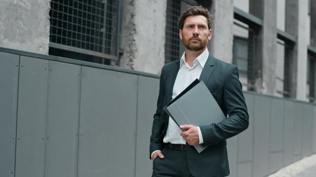 Confident serious Caucasian formal business man walking in city holding folder with documents going to office building in morning work day. Mature businessman male middle-aged leader walk go to agency