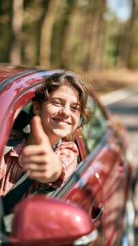 a young woman seated in a red car. She is smiling and giving a thumbsup with her right hand. She wears a red and white checkered shirt and a seatbelt.