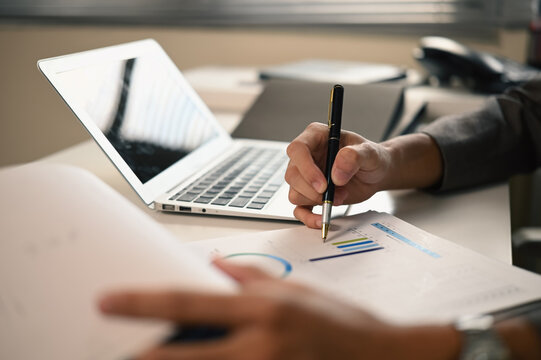 Cropped Hands of Business Person Analyzing Financial Charts with Pen and Laptop on Office Desk