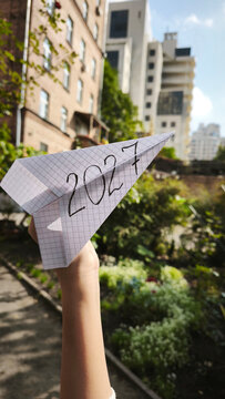 Paper airplane marked 2027 held above a modern city garden, symbolizing the future, urban development, new opportunities, dreams, ambition, and plans for the coming year.