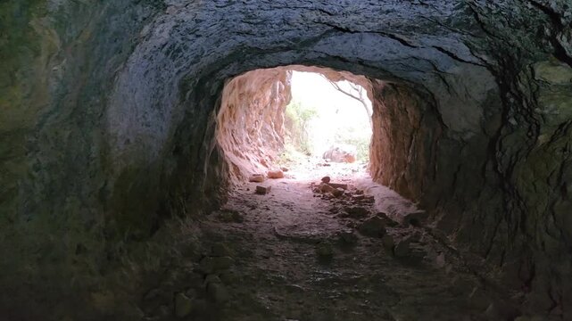 Walking out of dark rocky cave tunnel towards bright sunlight and green forest, POV shot.