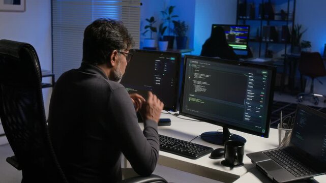 Middle aged South Asian programmer analyzing cybersecurity code on monitors in office. Visual asset perfect for IT infrastructure, software engineering, and data defense projects
