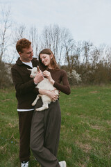 Naklejka premium Cheerful couple outdoors, holding a goat kid, smiling beneath the sun, casual attire, open field, soft breeze, natural colors, lush grass, warm connection, relaxed pose, candid moment, authentic