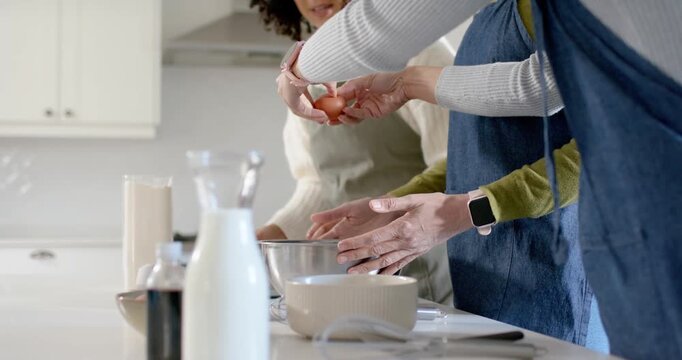 Two adult women passing eggs and cracking them into mixing bowl at kitchen island, preparing batter