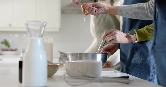 Two women cracking eggs, whisking mixture to prepare batter at kitchen island with bowl and whisk