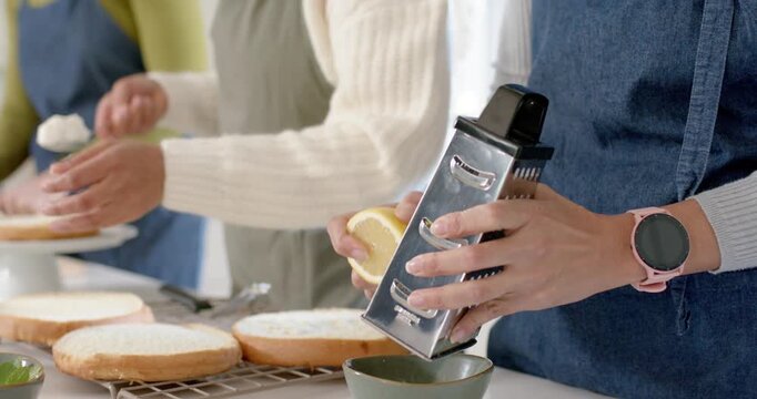 Right hands holding lemon and grater, zesting into green bowl while left hands smoothing cakes