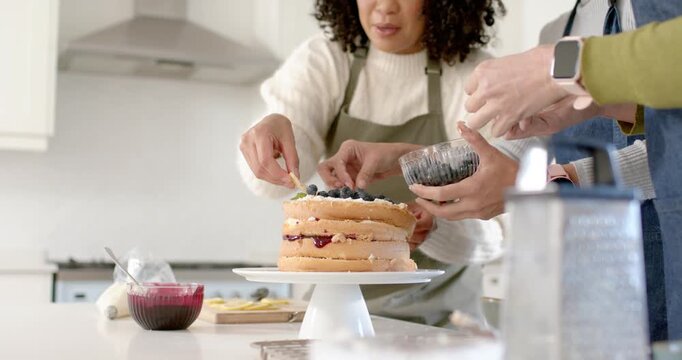African American women in aprons handing berries, adding final touches on 3-layer cake in kitchen