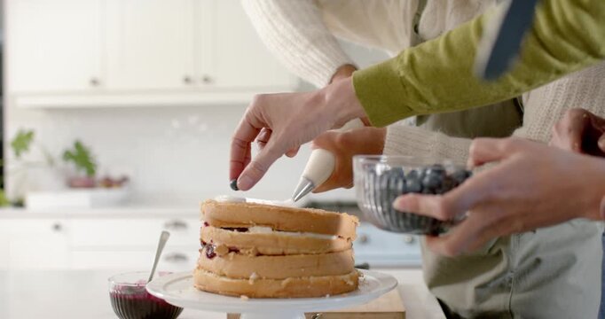After stacking, two bakers are placing blueberries on three-layer cake to decorate in kitchen