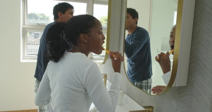Couple being prompted by morning light brushing teeth at sink with yellow brush, man adjusting hair