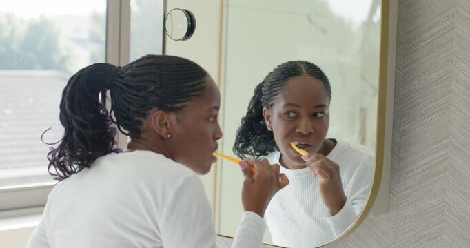 African American woman bringing yellow brush to mouth, brushing teeth while checking form at mirror