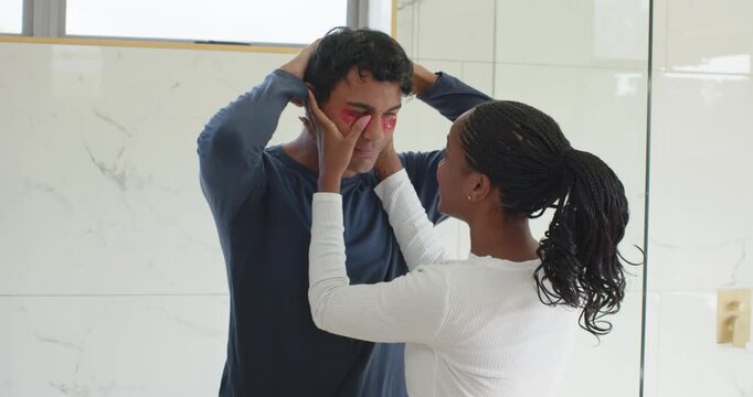 African American couple by mirror, with man holding red patches, woman applying them for eye care