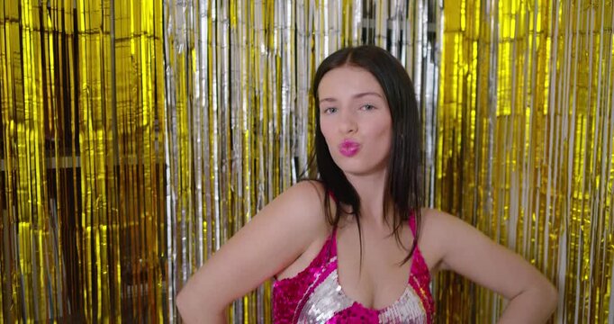 Woman wearing pink sequin dress and hoops, posing and blowing gold party horn with fringe backdrop