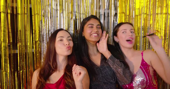 Diverse female friends prompted by gold fringe in sequin dresses posing in booth, using blowers