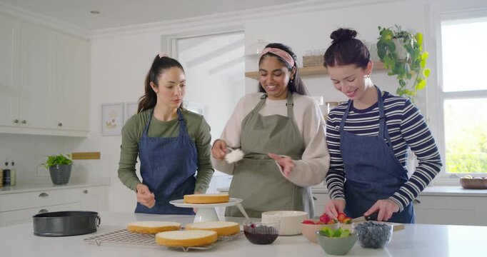 Diverse female friends placing cake layer on turntable stacking and frosting cake at kitchen island
