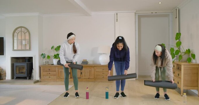 Diverse female friends placing bottles, unrolling mats at home, warming up, closing palms for yoga