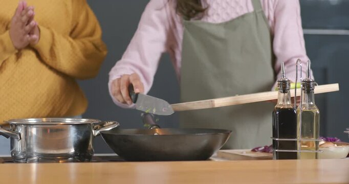 Female friends tipping cutting board and scraping veggies with cleaver into pan at counter cooking