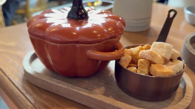 Closeup of ceramic orange pumpkin soup cocotte bowl and small metal dish with white bread croutons served on wooden tray in restaurant. Traditional autumn seasonal soup serving,  culinary food concept