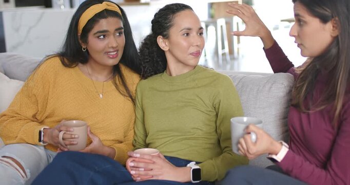 Diverse female friends holding mugs on sofa as right woman sharing story, gesturing and smiling