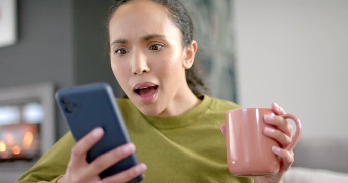 African American woman on couch holding phone and mug, glancing, reacting then refocusing to screen