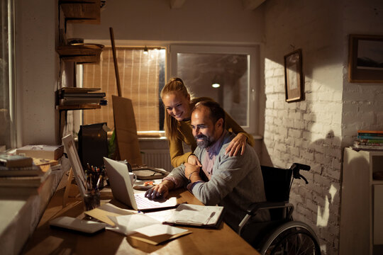 Woman hugs wheelchair user as he works on laptop at home