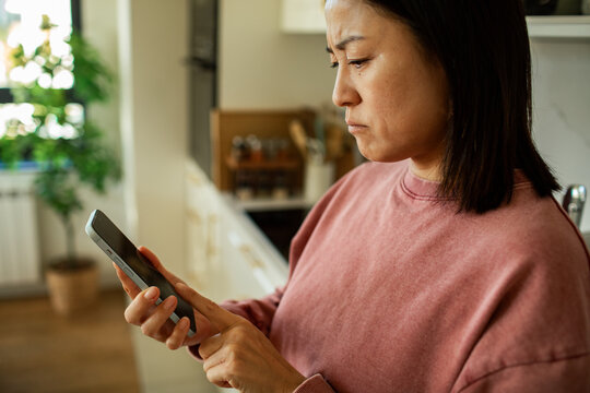 Woman frowning at smartphone while texting in home kitchen