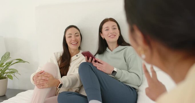 Diverse women friends sitting on white bed, responding to gesturing woman while scrolling red phone