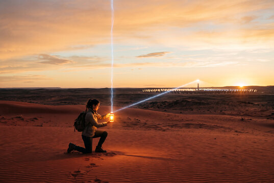 Desert explorer kneels at sunset with lantern and light beam