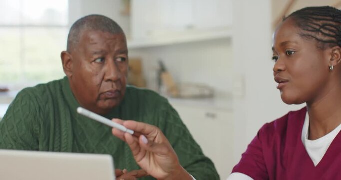 African-American nurse leaning in while guiding senior using laptop at kitchen table, pointing pen