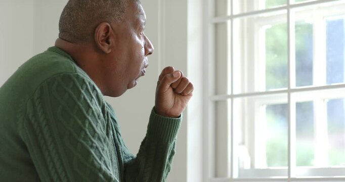 African American man sitting by window briefly coughing into fist, hand on chest calming