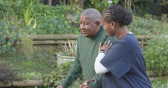 Carer in scrubs placing hand on shoulder guiding cane-walking client through garden steadying steps