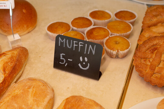 Close-up of plain muffins in white paper cups arranged on bakery display counter with nearby bread and small chalkboard price sign reading muffin.