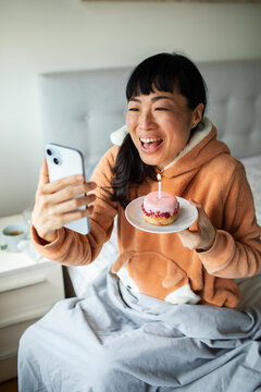 Woman on video call holding birthday cupcake in bed
