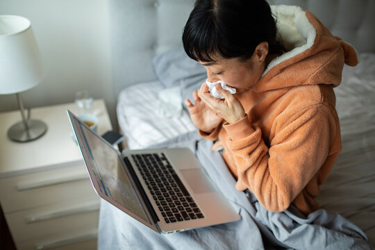 Sick woman blowing nose while using laptop in bed