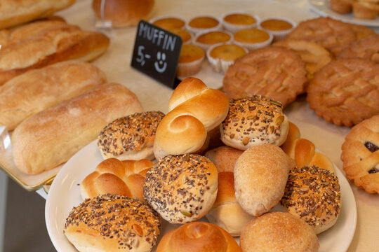 Close view of assorted bread rolls and seeded buns arranged on white serving platter, with additional loaves and muffins visible in background.