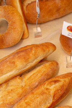Close-up view of several freshly baked baguettes with golden crusts displayed beside sesame seeded bagel on bakery counter, with small sign holders visible in background.