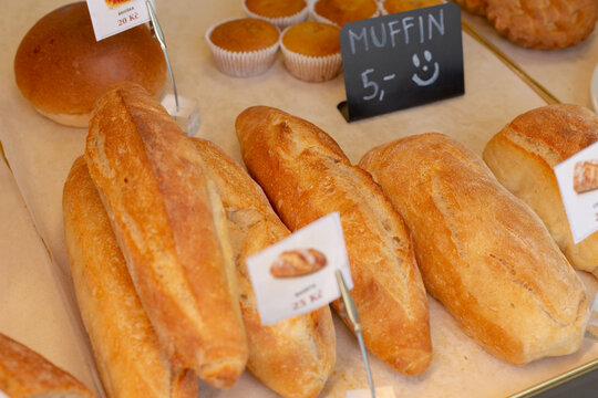 Close view of freshly baked baguettes and round rolls on paper-lined bakery counter, with chalkboard sign reading muffin and muffin cups in soft background.